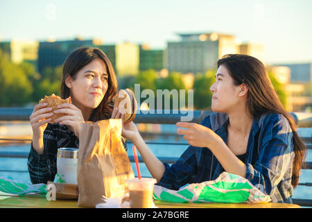 Zwei biracial Asiatische, Kaukasische jugendlich Mädchen oder Frauen youmg Essen Hamburger im Freien bei Sonnenuntergang am See im städtischen Bereich, reden und lächeln Stockfoto