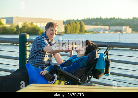 Kaukasische Vater, der in den frühen 50er Jahren 13 Jahre alten Sohn im Rollstuhl einen Hamburger im Freien an einem sonnigen Tag Fütterung von Lake Park im städtischen Bereich deaktiviert Stockfoto