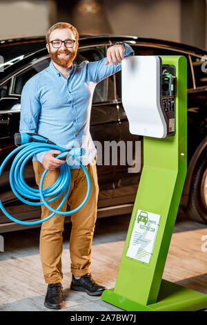 Voller Körper Porträt eines jungen Mannes mit Ladekabel in der Nähe der Batterieladestation im Autohaus. Konzept der Schnellen home car Ladegeräte Stockfoto