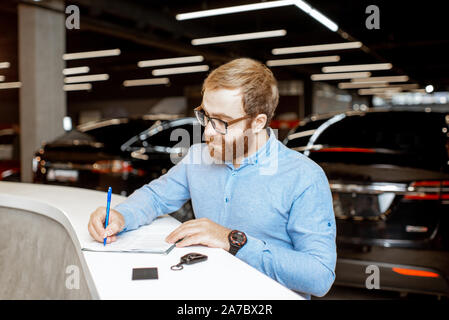 Junger Mann Unterzeichnung einige Dokumente, den Kauf oder die Anmietung eines Autos in der modernen Showroom des Autohauses Stockfoto