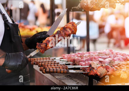 Koch braten Fleisch vom Grill. Koch in schwarze Schürze frites Fleisch auf Feuer. Grill im Freien. Helles Sonnenlicht bokeh Stockfoto