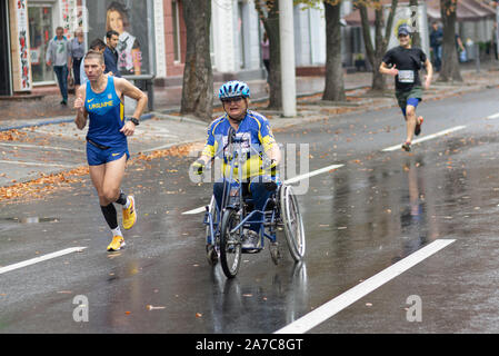 DNIPRO, UKRAINE - September 16, 2018: ältere Frau, die auf einem Stuhl neben normalen Athleten an einem regnerischen city street während 10 km Abschnitt eines Stockfoto