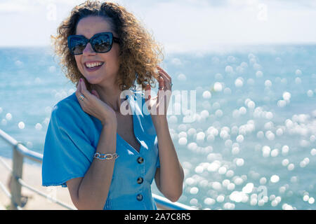 Touristische Frau im Sommerkleid und Sonnenbrille lächelnd und mit dem blauen Mittelmeer auf dem Hintergrund glücklich Stockfoto