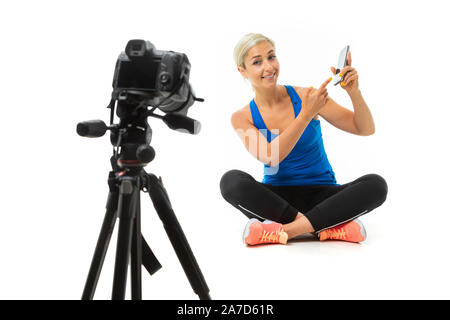 Der junge sportliche Mädchen mit blondem Haar sitzt vor der Kamera und zeigt Telefon Stockfoto