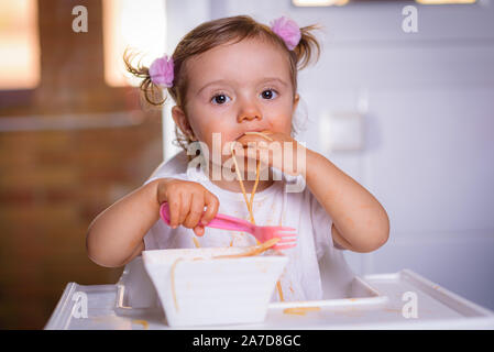 Süße kleine Funny Girl von 18 Monate / 1 Jahr alt, Baby, Mädchen, in einer leistungsstarken Stuhl zu Hause sitzt, isst Nudeln mit Löffel und Gabel, Foto innen Stockfoto