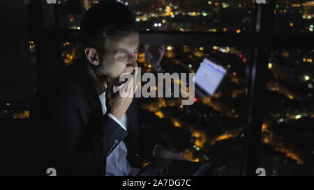 Sleepy junger Mann in Anzug arbeitet auf seinem Computer in der Nacht in der Nähe der Fenster mit Panoramablick auf die Stadt. Er nimmt seine Brille ab und rieb müde Augen. Überstunden Konzept. Stockfoto
