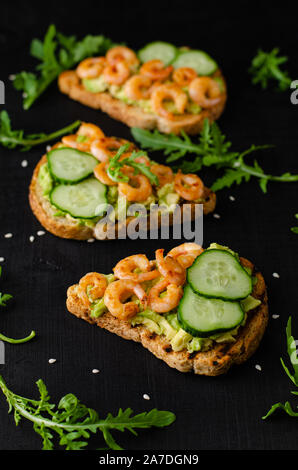 Leckere Toasts mit gebratenen Garnelen, Avocado und Gurke auf schwarzen Hintergrund. Vertikale, selektiver Fokus Stockfoto