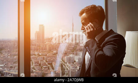 Junge erfolgreiche Unternehmer in Bürosoftware reden ist Mobiltelefon, Seitenansicht. Er steht in der Nähe der Fenster mit Panoramablick auf die Stadt in seinem Büro an die Arbeit. Stockfoto