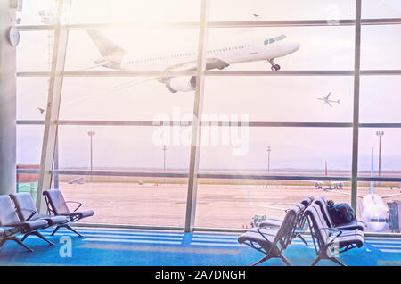 Blick auf die sonnenbeschienenen Fliegen Flugzeug über Flughafen Fenster Tor. Flughafen abreise Sitzgelegenheiten in der Lounge mit Blick auf den fliegenden Flugzeug Stockfoto