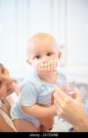 Das Konzept der Mutterschaft, Nanny, Kindheit und Kindheit. Innen- schuss in der Küche. Zwei Frauen und ein Kind in ihren Armen, das Kind Wasser gegeben wird, ein Stockfoto