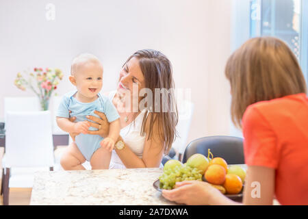 Das Konzept der Mutterschaft, Nanny, Kindheit und Kindheit. Innen- schuss in der Küche. Zwei Frauen und ein Kind in ihren Armen, das Kind Obst angeboten wird, Stockfoto