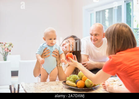 Das Konzept der Mutterschaft, Nanny, Kindheit und Kindheit. Innen- schuss in der Küche. Zwei Frauen und ein Kind in ihren Armen, das Kind Obst angeboten wird, Stockfoto