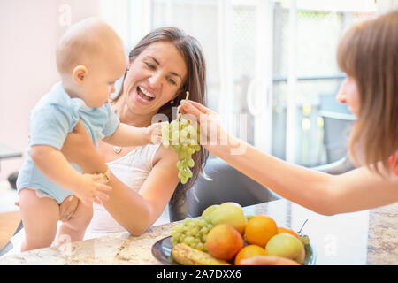 Das Konzept der Mutterschaft, Nanny, Kindheit und Kindheit. Innen- schuss in der Küche. Zwei Frauen und ein Kind in ihren Armen, das Kind Obst angeboten wird, Stockfoto