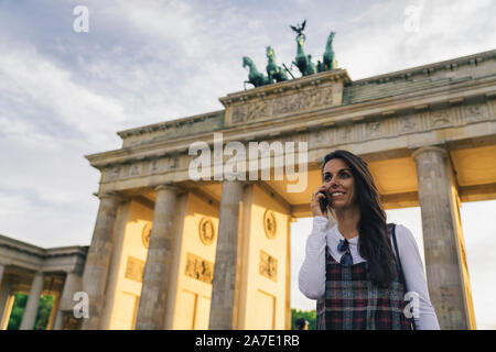Portrait von lächelnden jungen Frau im Gespräch über ein Handy gegen Brandenburg Schicksal in Berlin, Deutschland. Stockfoto