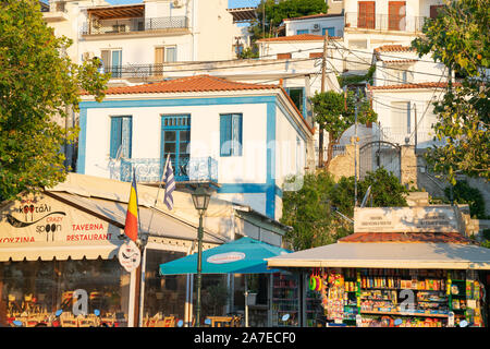 SKIATHOS GRIECHENLAND - 30. JULI 2019; Geschäfte und Restaurants mit tioered Unterkunft und Gehäuse hinter entlang der Waterfront Stockfoto