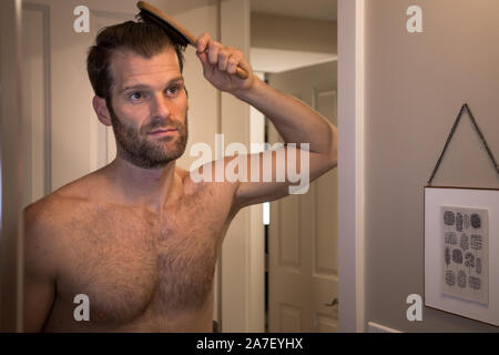 Hübscher junger Kaukasusmann, der sich in einem Badezimmerspiegel ansieht, während er seine Haare kämmt. Stockfoto