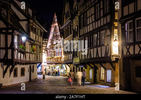 Menschen flanieren in der Nacht in einer gepflasterten Straße des Viertels Petite France in Straßburg, Frankreich, gesäumt von Fachwerkhäusern und Restaurants. Stockfoto