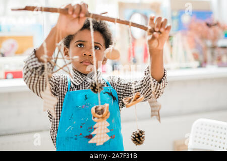 Cute ernsthafte Schüler in der blauen Schürze Stick mit handgefertigten Weihnachten Spielzeug suchen Stockfoto