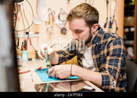 Ernsthafte Handwerker biegen über gebrochene Gadget auf dem Tisch, während mit einem Schraubendreher Stockfoto