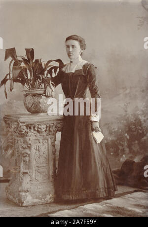 Späten viktorianischen oder frühen Edwardian Kabinett Foto Karte Porträt der jungen Edwardian Frau mit einem Buchstaben und tragen eine düstere, dunkle Kleidung, neben Topfpflanze posing - eine aspidistra, waren beliebte Pflanzen in dieser Zeit. ca. 1901, Truro, Cornwall, Großbritannien Stockfoto