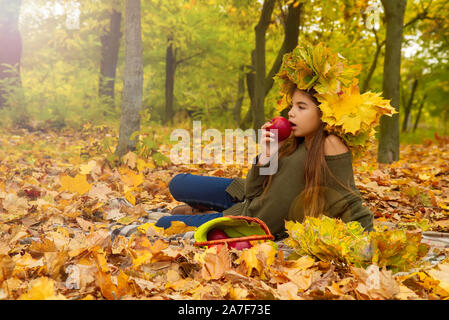 Kleine Mädchen bei einem Picknick im Herbst Park isst ein reifer roter Apfel sitzt auf gefallene Laub in einem Kopfschmuck aus Ahorn Blätter Stockfoto