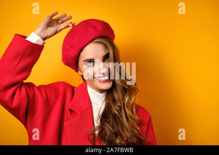 Ein Mädchen mit Locken, roten Lippenstift in einem roten Mantel steht auf einem gelben Hintergrund und lächelnd Zieht die Finger nach oben. Stockfoto