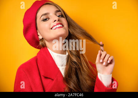 Ein Mädchen mit Locken, roten Lippenstift in einem roten Mantel und eine Baskenmütze steht auf einem gelben Hintergrund, twirling a mit Ihrem Finger curl. Stockfoto