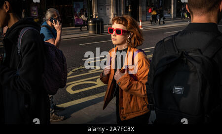 Eine junge Frau macht, an der Londoner Oxford Street mit der tief stehenden Sonne Kommissionierung ihre rote Sonnenbrille durch die Menge Stockfoto