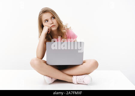 Uninterested cute blonde Junge Kind Mädchen, Sitzen mit gekreuzten Beinen laptop, schlanken Kopf auf Gesicht und Gelangweilt vom Computer Bildschirm schauen, hassen Stockfoto