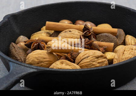 Walnüsse, Haselnüsse, Paranüsse und Walnüsse in einer gusseisernen Pfanne mit Zimtstangen und Sternanis. Auf einem grauen Tischdecke Stockfoto