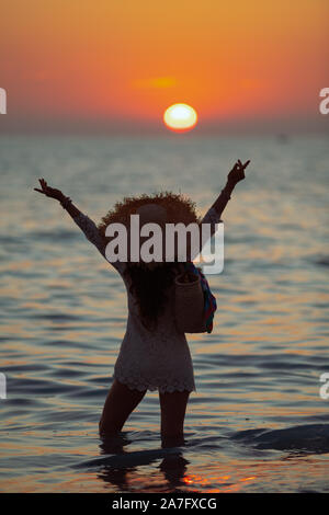 Silhouette der modernen Frau in weißem Kleid auf dem Strand bei Sonnenuntergang im Meer und Jubel. Stockfoto