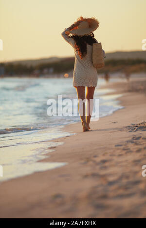 In voller Länge Porträt der 40 Jahre alte Frau in weißem Kleid und Strohhut am Strand bei Sonnenuntergang wandern. Stockfoto