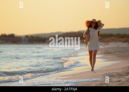 In voller Länge Porträt der elegante 40-jährige Frau in weißem Kleid und Strohhut am Meer bei Sonnenuntergang wandern. Stockfoto
