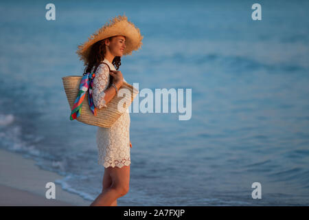 Seitenansicht des nachdenklich moderne Frau in weißem Kleid und Strohhut auf dem Ozean Küste bei Sonnenuntergang. Stockfoto