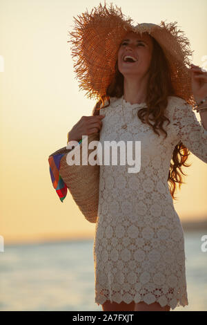 Lächelnd 40 Jahre alte Frau in weißem Kleid und Strohhut am Strand bei Sonnenuntergang Spaß Zeit. Stockfoto