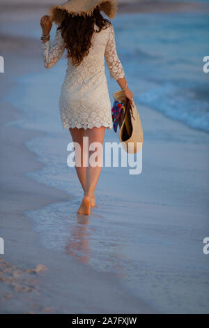 Aus hinter der modernen 40-jährige Frau in weißem Kleid und Stroh Hut auf dem Strand bei Sonnenuntergang wandern. Stockfoto