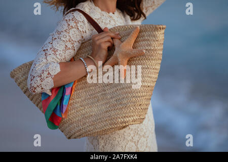 Nahaufnahme auf stilvolle Frau in weißem Kleid auf die Küste bei Sonnenuntergang, Strand Stroh Beutel und Seesterne. Stockfoto