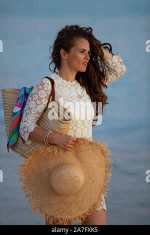 Moderne mittlere Alter Frau in weißem Kleid und Strohhut am Meer bei Sonnenuntergang der Blick in die Ferne. Stockfoto
