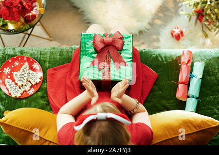 Urlaubszeit. Obere Ansicht von lächelnden Moderne kleine Prinzessin im roten Kleid mit Weihnachtsgeschenk, Klatschen, beim Sitzen auf der Couch in der modernen Liv Stockfoto