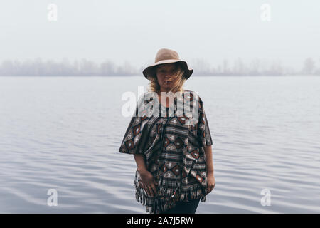 Frau mit Hut und Poncho stehen am Ufer des Flusses und suchen zum Herbst nebligen Wald. Stockfoto