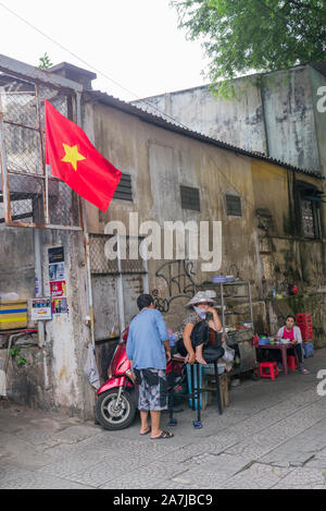 Saigon Seitenszene mit leuchtend roter Vietnam-Flagge und lokalen Straßenverkäufern, die Roller parken, die Frau, die lächelnd auf dem Stuhl sitzt und die Kamera betrachtet. Stockfoto