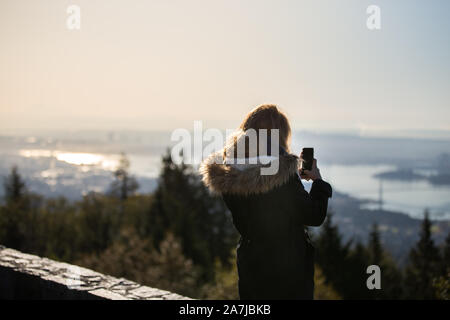 Eine junge Frau in einen Wintermantel am Cypress Mountain Lookout ein Foto von Downtown Vancouver auf Ihr Handy. Stockfoto