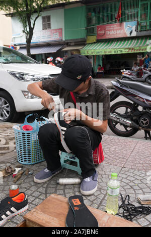 Ein junger Vietnamese, der eine schwarze Baseballkappe trägt, sitzt auf Holzhocker auf dem Gehweg und repariert ein Paar schwarzer Sneaker. Stockfoto