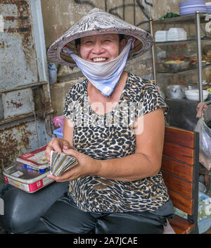 Lächelnde, vietnamesische Straßenverkäuferin, die Strohhut trägt, weißer Kinnschal auf dem Stuhl sitzt und Geld einen großen Stapel auf Geld in der rechten Hand zählt Stockfoto