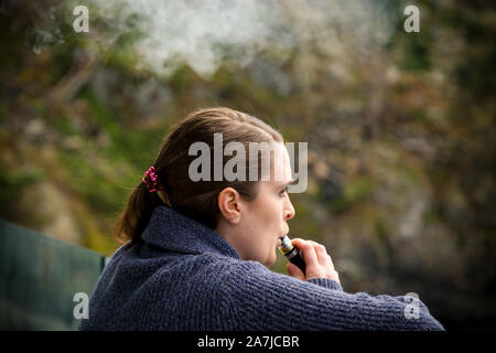 Junge brunette kaukasische Frau rauchen eine vape ecigarette beim Stehen auf einem Balkon mit Blick auf den Ozean. Stockfoto