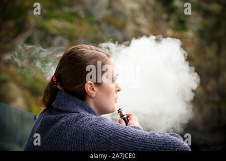Junge brunette kaukasische Frau rauchen eine vape ecigarette beim Stehen auf einem Balkon mit Blick auf den Ozean. Stockfoto