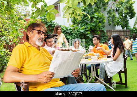 Senior Großvater lesen Zeitung mit Familie mit Frühstück in den Hintergrund - Innenhof in der Nähe Ihres Hauses Stockfoto