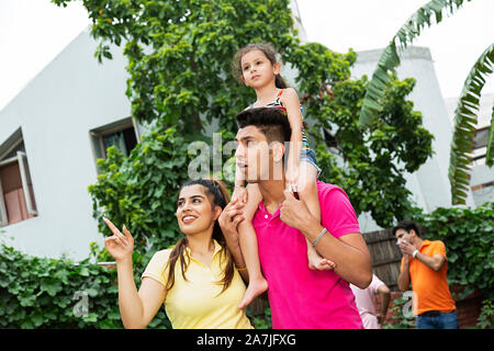 Glückliche junge Familie Mutter mit Kind Mädchen auf Schultern und Papa zeigen auf etwas im Hof in der Nähe von Ihrem Haus Stockfoto