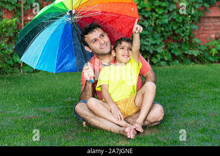 Fröhlich-Familie Vater und Kind Junge sitzt im Gras mit Schirm zeigte auf etwas in - der Regen im Garten Stockfoto
