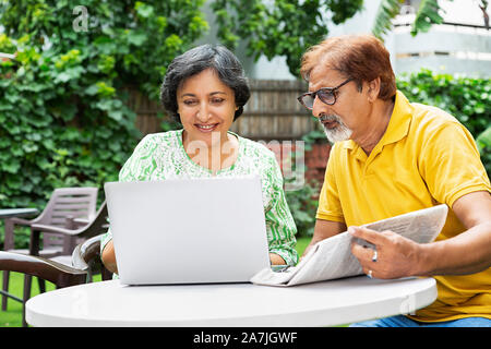 Indische ältere Paare männlich und weiblich auf der Suche - auf Laptop Bildschirm mit Zeitung sitzen im Innenhof Ihres Hauses Stockfoto